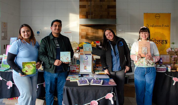Cuatro personas (tres mujeres, un hombre) sonriendo detrás de una mesa llena de libros y decorada con una guirnalda de setas en un evento comunitario de Gota de Lluvia. La mujer de la izquierda sostiene un libro verde titulado "Dulce y Suave", el hombre sostiene el libro "nido", y la mujer de la derecha sostiene el libro "Dulce Pincelada de mi nostalgia". La mesa tiene otros carteles, incluido uno para té, infusiones e hamsa, y un cartel central de un libro ilustrado.