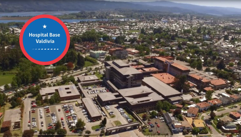 Vista aérea del Hospital Base Valdivia en Chile, mostrando su gran complejo de edificios y estacionamientos rodeado por la ciudad y montañas.