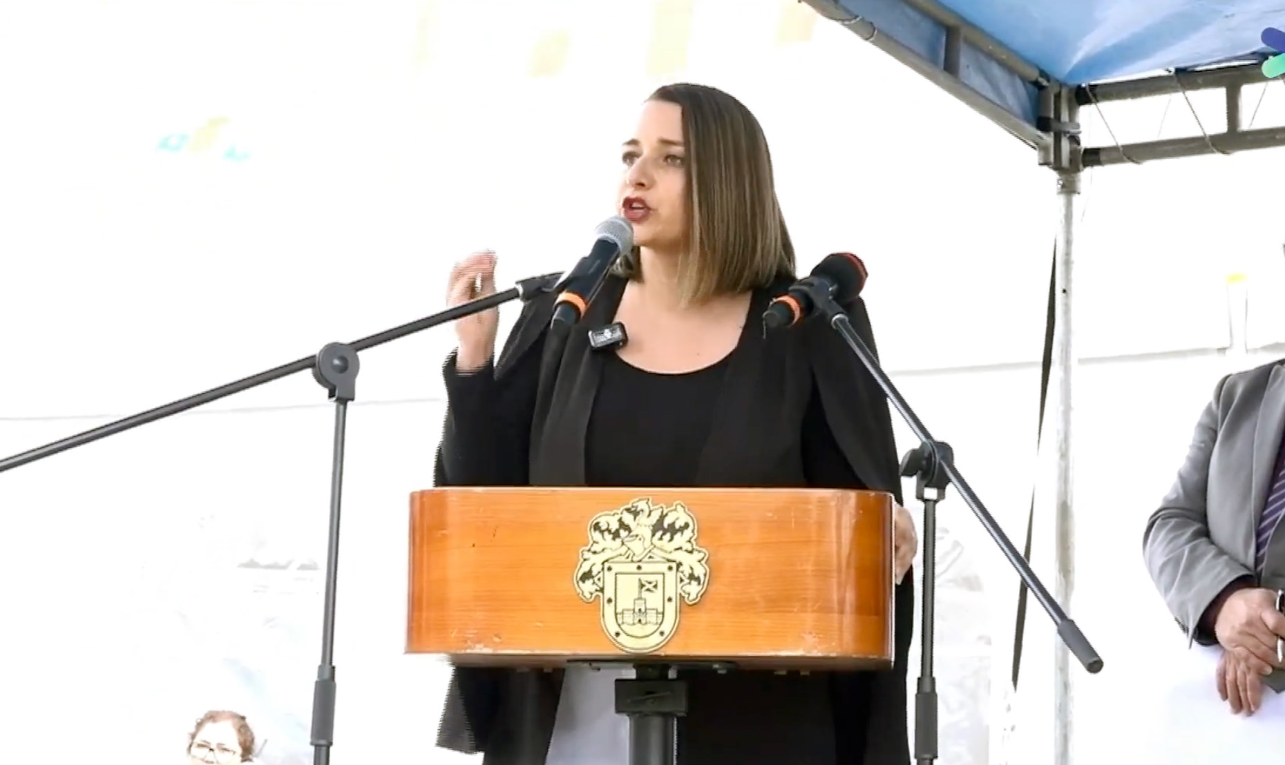 Carla Amtmann, alcaldesa de Valdivia, de pie detrás de un podio de madera con el escudo de armas dorado de la ciudad, hablando apasionadamente en un micrófono con gestos de la mano.