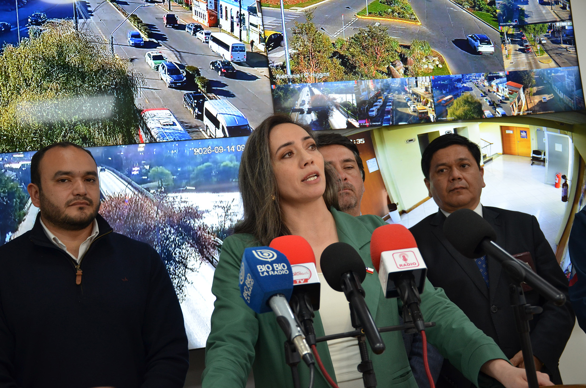 Una mujer con una chaqueta verde de traje habla en una rueda de prensa con micrófonos de "BIO BIO LA RADIO" e "I RADIO" delante, flanqueada por dos hombres con trajes oscuros. Detrás de ellos, una gran pantalla curva muestra vistas de cámaras de seguridad de tráfico en directo con marcas de agua de fecha y hora que indican el 14 de septiembre de 2026 a las 09:35.
