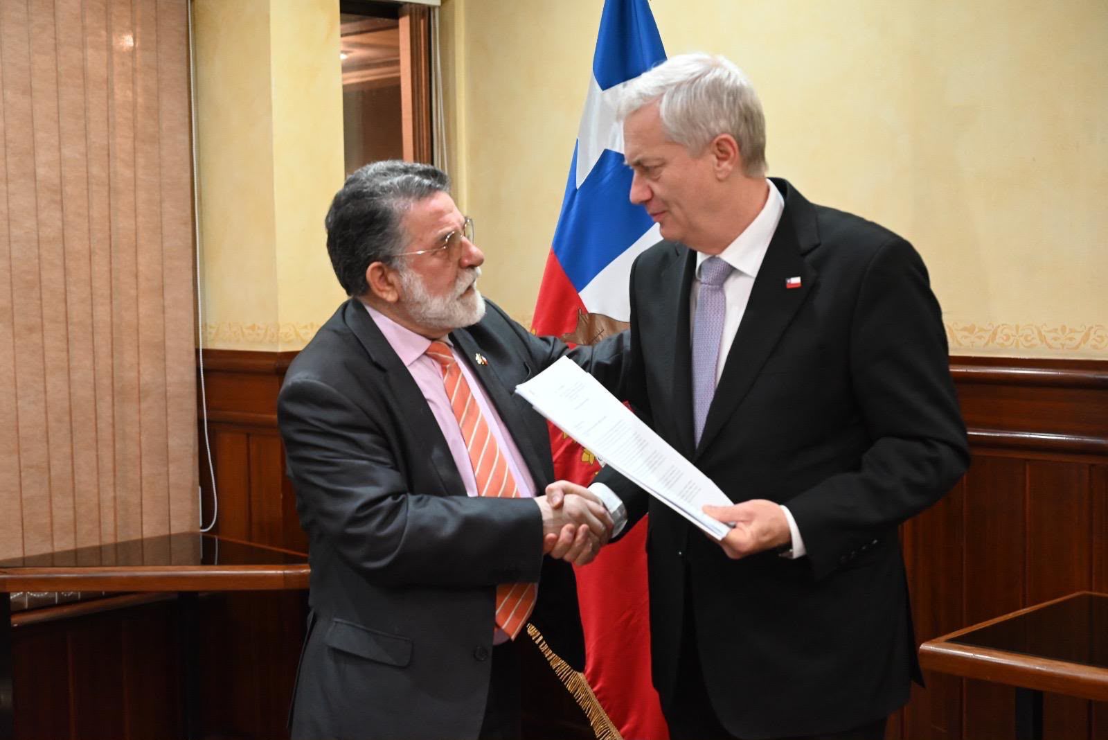 El Gobernador Regional de Los Ríos, Luis Cuvertino, estrecha la mano del Presidente José Antonio Kast, quien sostiene un documento de varias páginas. Ambos hombres están de pie, vestidos de traje, frente a la bandera oficial de la Región de Los Ríos y paneles de madera decorativos.