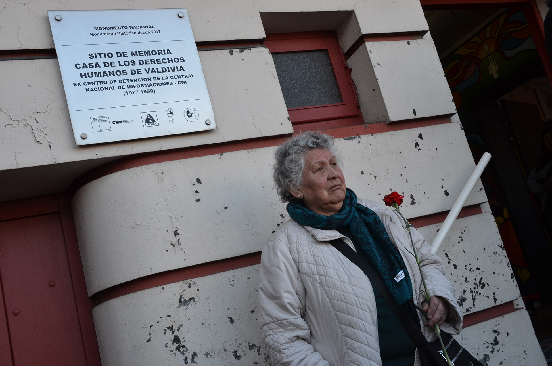 Una mujer mayor con cabello canoso rizado y bufanda azul mira a la derecha, sosteniendo un clavel rojo y una vara blanca. Está frente a una pared con una placa de metal que dice: "SITIO DE MEMORIA CASA DE LOS DERECHOS HUMANOS DE VALDIVIA. EX CENTRO DE DETENCION DE LA CENTRAL NACIONAL DE INFORMACIONES - CNI (1977 1990)".
