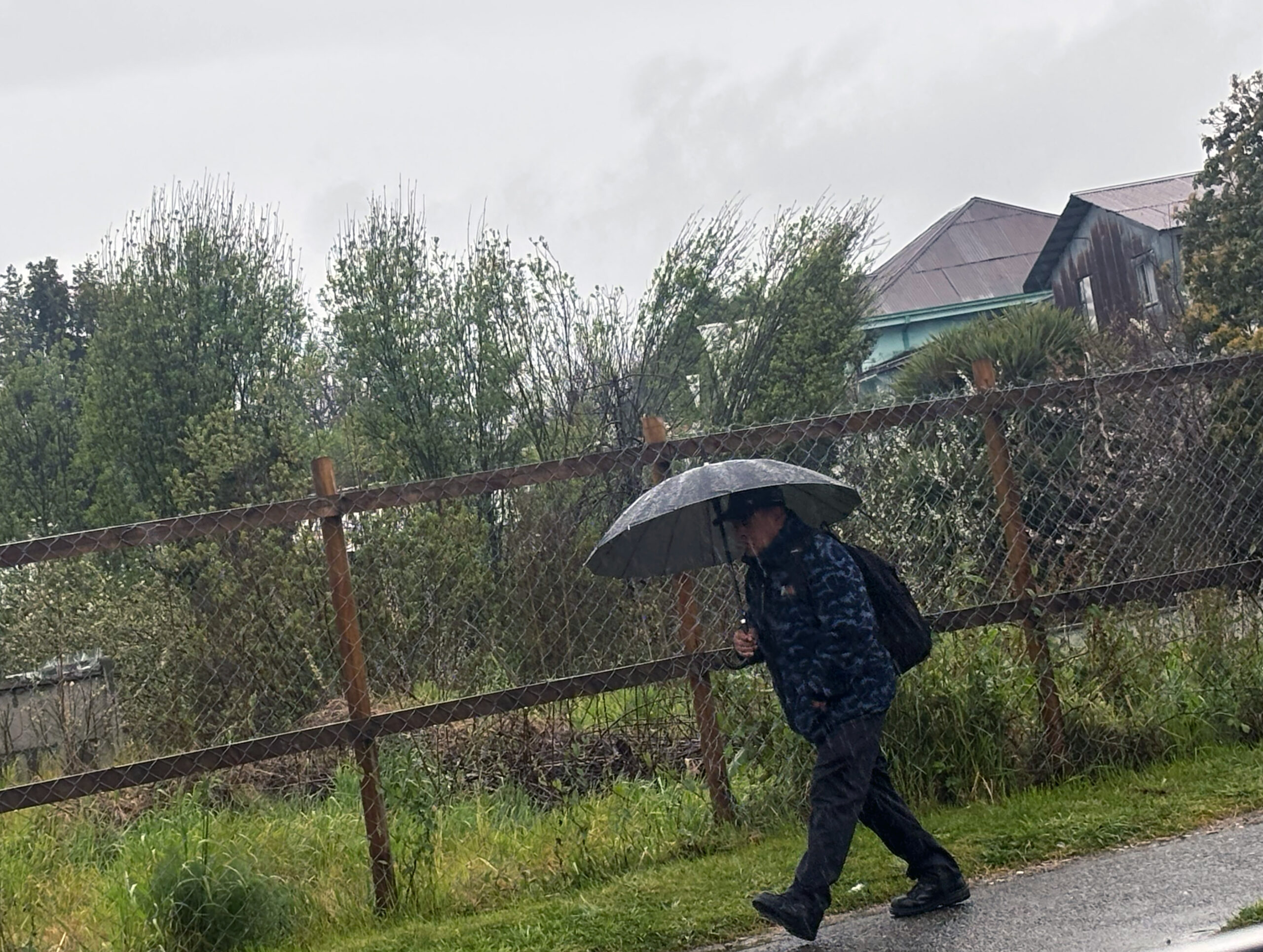 Persona caminando bajo la lluvia con un paraguas abierto y mochila junto a un cerco de malla