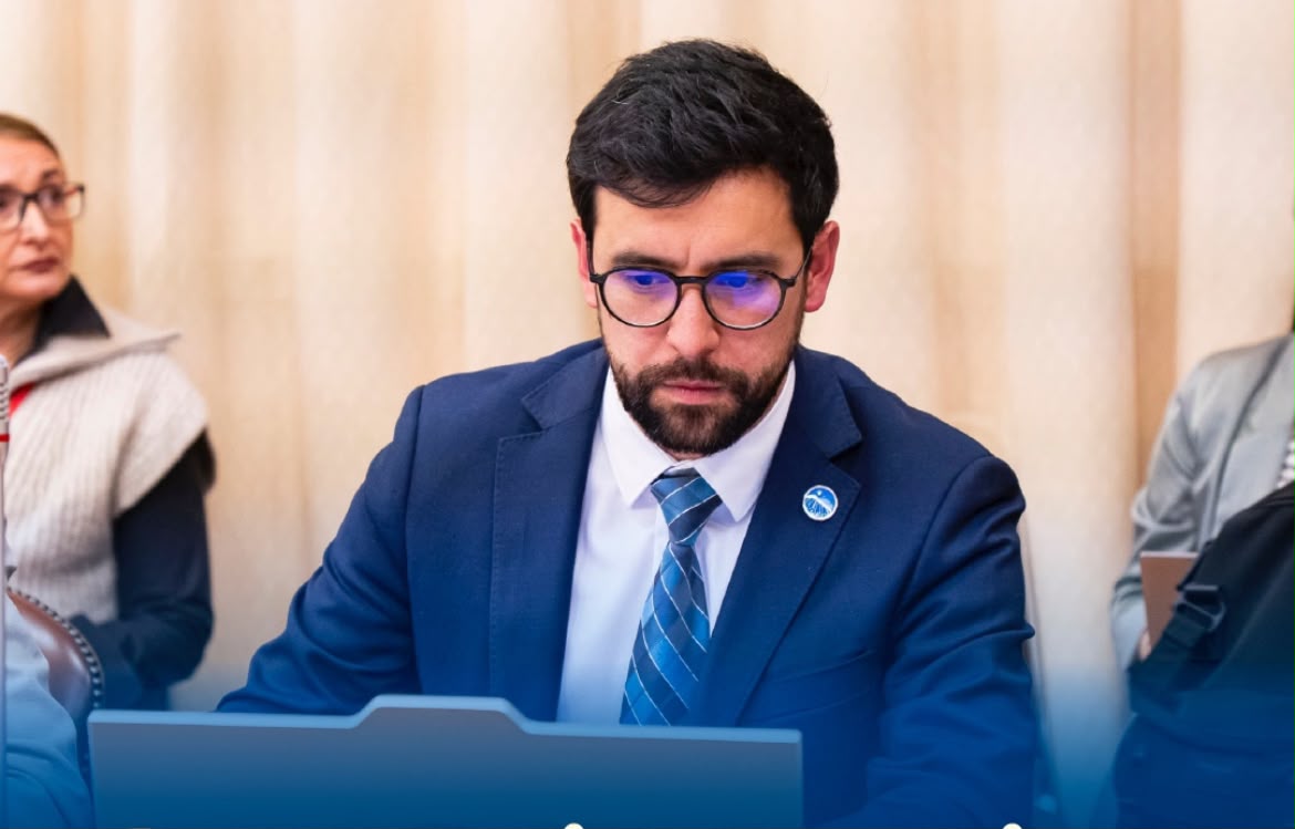 Hombre de traje azul, anteojos y barba mirando hacia abajo durante una reunión