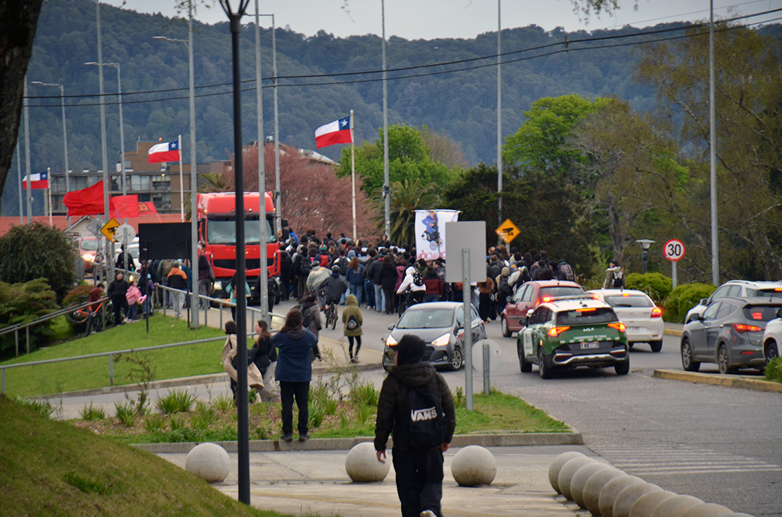 Multitud marchando en una avenida con banderas de Chile, acompañada por un camión rojo y una patrulla de Carabineros